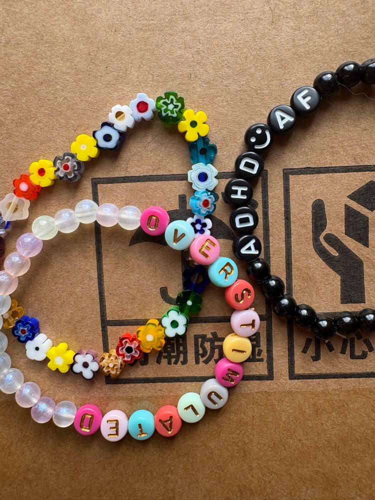 Bracelets with letter beads. One says OVERSTIMULATED and the other says ADHD AF. Another is made of flowers beads