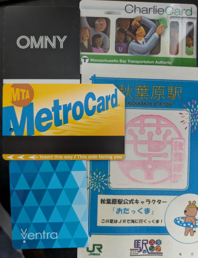 Various public transit cards and stamps on a black table arranged in a grid. Clockwise starting from top left: NYC OMNY, MBTA CharlieCard, an Eki stamp for JR-East Akihabara Station in Tokyo, Chicago Ventra, and NYC MetroCard