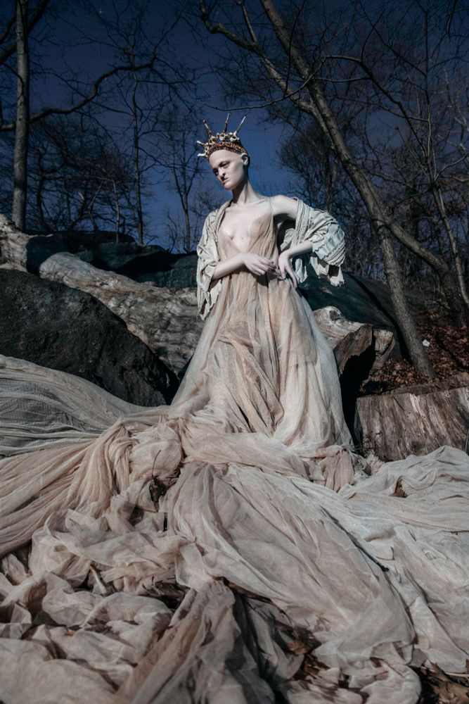 Model Melanie Gaydos stands in a clearing photographed by Ekaterina Belinskaya. Melanie is wearing a dramatic tea stained cotton tulle gown draped over the rocks along with a headpiece of copper wrapped deer bone, both by Corvus Crux