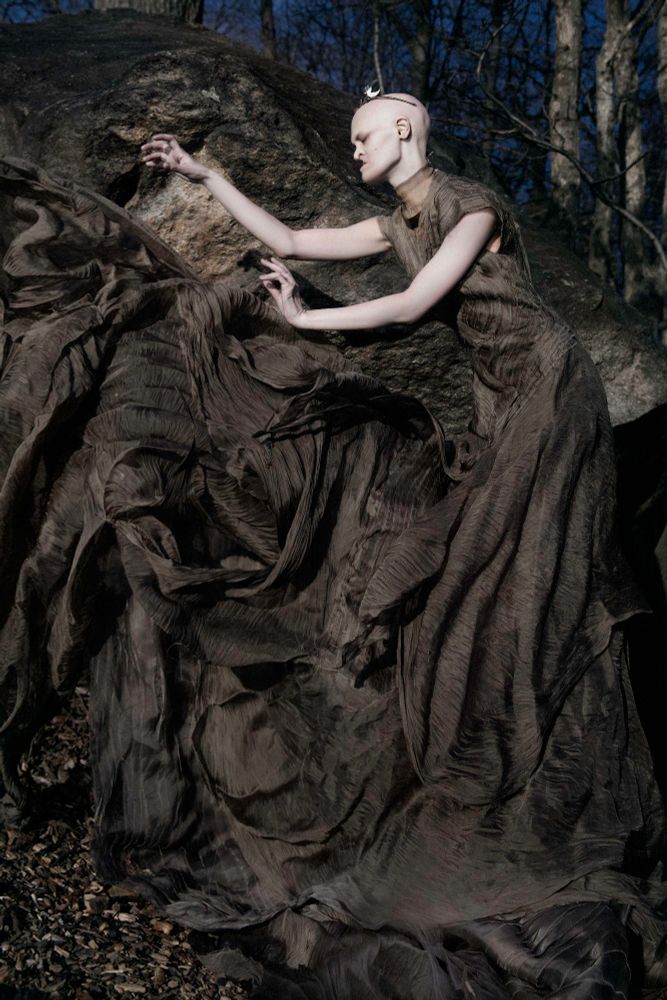 Model Melanie Gaydos reaches out to something  unseen in a clearing photographed by Ekaterina Belinskaya. Melanie is wearing a pleated and hand woven toffee colored silk gown by Corvus Crux
