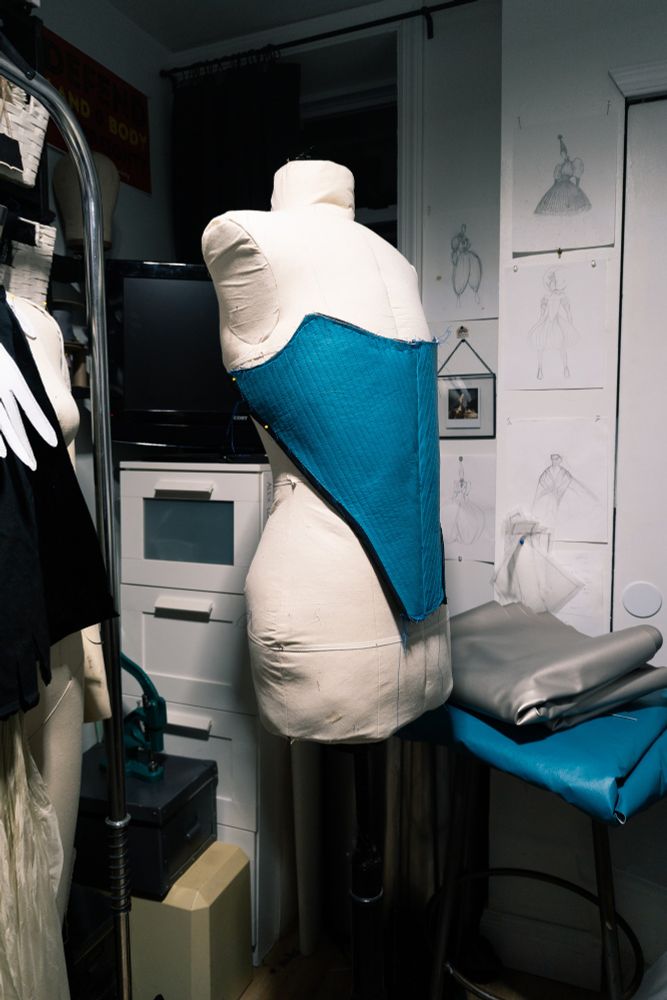 Work in progress photo of blue 16th century inspired stays on custom dress form for Tillie the Clown in small studio. Sketches of ongoing  work on back wall.