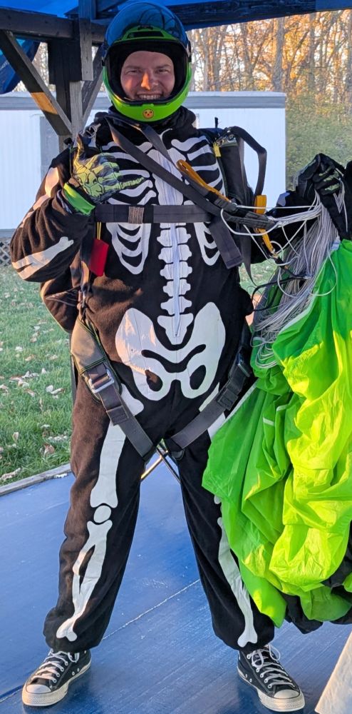A white guy wearing a skeleton onesie halloween costume, black and white converse shoes, skeleton gloves and lime green helmet. His visor is up and he's smiling. He is holding a lime green parachute and lines in one hand and giving a shacka with the other. 