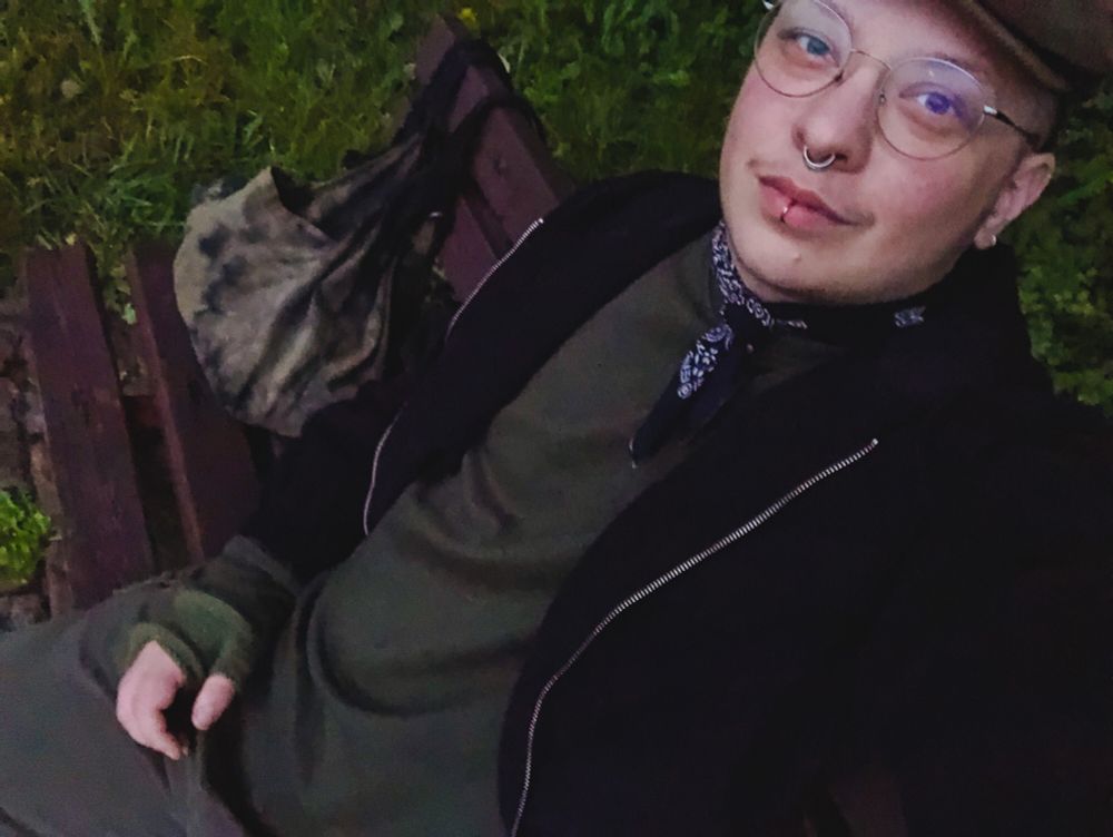 Jay sitzt auf einer Bank. Sichtbar sind der grüne Pullover, Hose, Armstulpen und Mütze und die schwarze Jacke und Halsband.