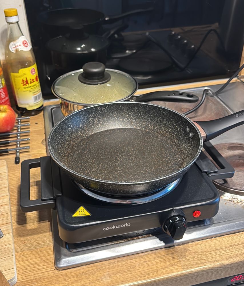 Hobplate single portable hob on top of an existing hob (broken). There is an empty frying pan atop it; with a filled saucepan behind. In the surroundings there is a wooden chopping board, trivet, Chinese vinegar, an apple. 