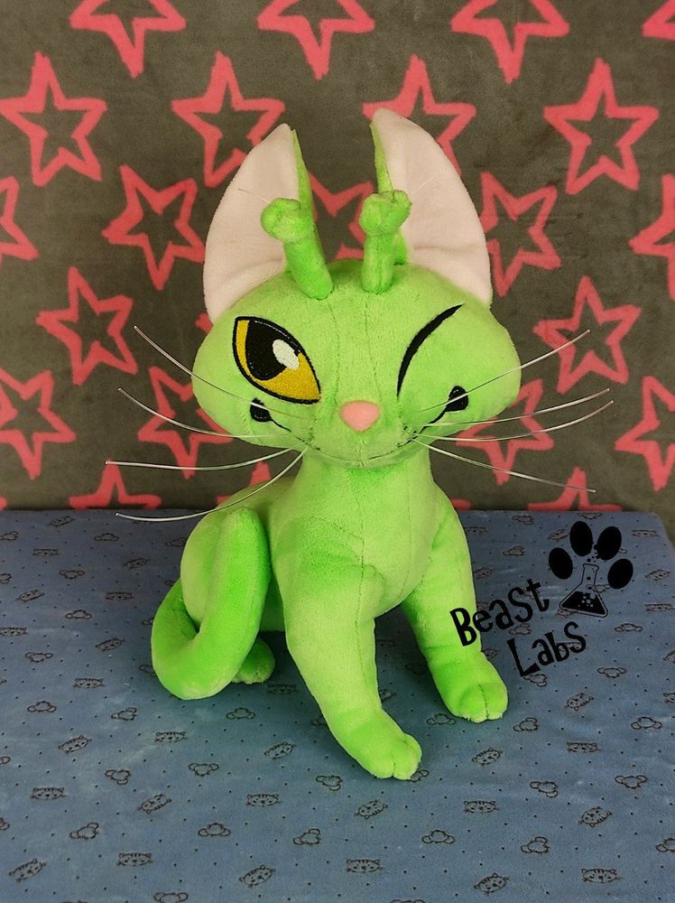 A green alien cat plushie. It is sitting on all fours, smiling, and winking.