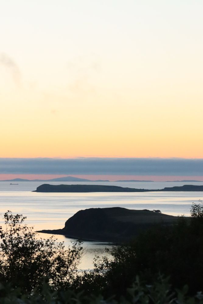 Looking from Skye to the Outer Hebrides. The sun has set, but the sky is still pale blue and yellow, the light late evening sky of summer in Scotland. The water is still and there is a low drift of blue cloud in the distance, near the horizon.