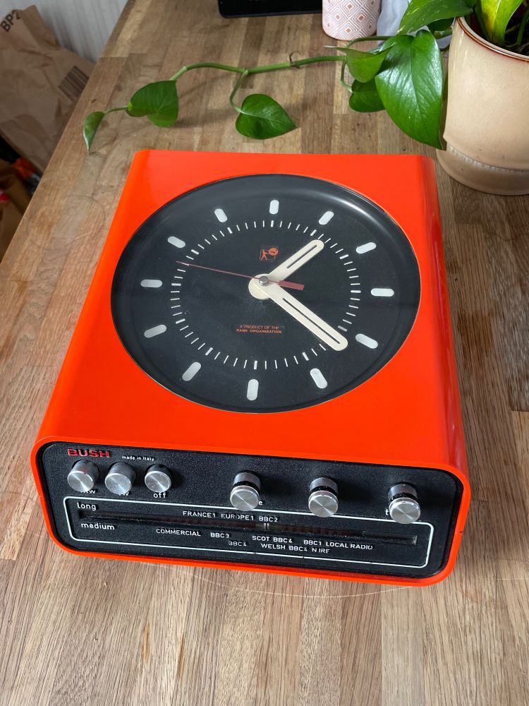 A bright orange clock radio, with a black face with white hands and hour markers. The radio part is inlaid in the base, with silver brushed steel knobs. The brand of the clock says ‘BUSH’ in the top left corner and along the bottom is the display, where it says long or medium which can be alternated with the knobs and a list of radio stations from BBC to Commercial and Local.