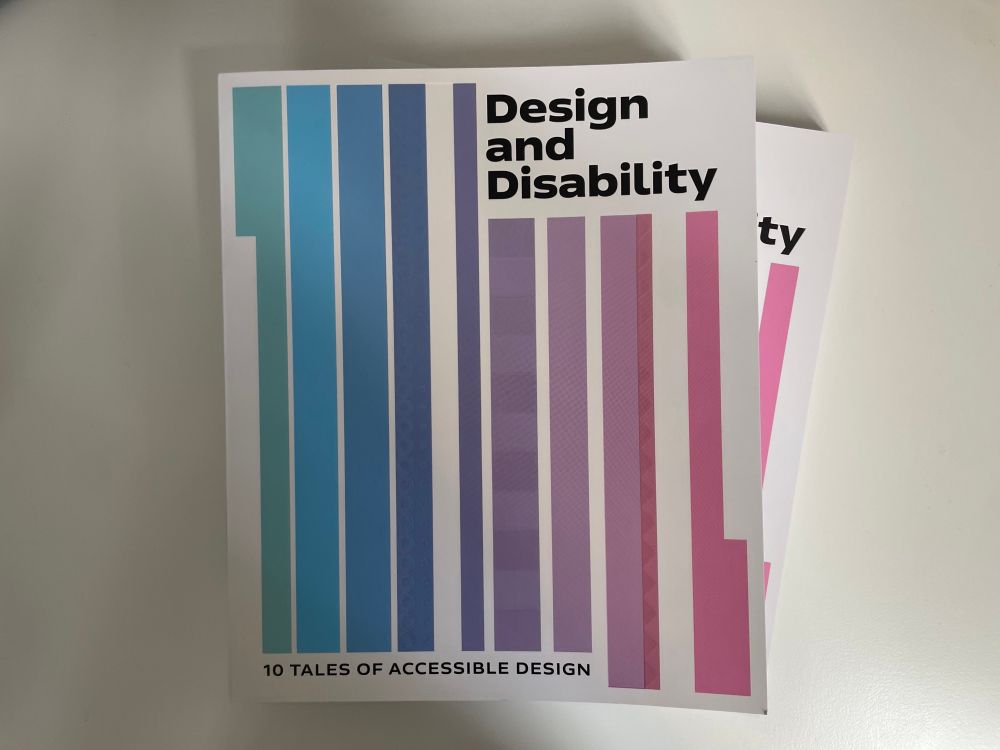 A book stacked on top of another on a table. The book cover is predominantly white. In the top right hand corner is the main title text in bold sans serif black font, reading Design and Disability. At the bottom left it reads in capitals, 10 Tales of Accessible Design. Across the front cover on a white background, from left to right is a series of long stripes of differing widths that go from top to bottom that go from each edge, with staggered gaps in between. They are in a colour spectrum starting from turquoise, to blue, to mauve, to purple to pink.