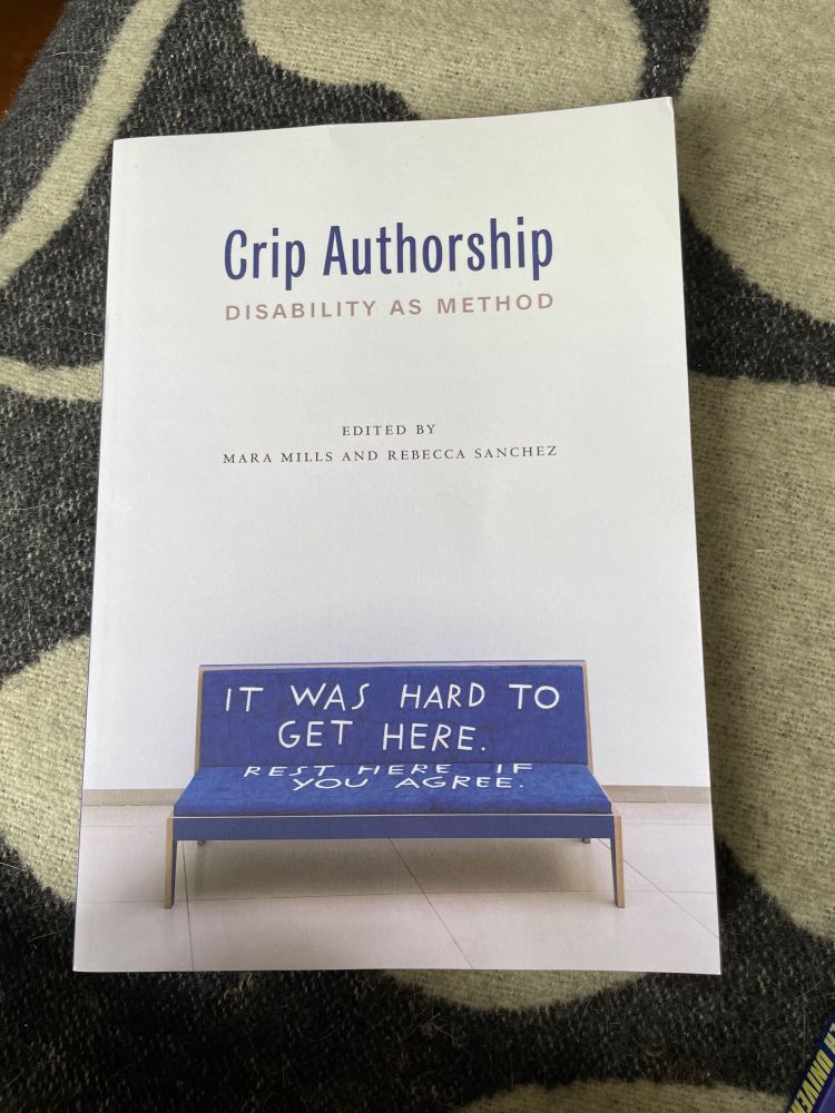 A book lays on a black and white abstract blanket. The book cover is of a white gallery space, with white walls and light grey tiled floors. A blue velvet cushioned bench is embellished with big white capitalised letters ‘It was hard to get here. Rest here if you agree.’  The book’s title is Crip Authorship: Disability as Method. Edited by Mara Mills and Rebecca Sanchez.