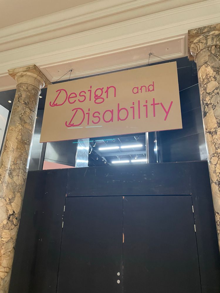 A large wooden sign hangs between two pale marble columns above two closed black doors. On the sign reads in fuschia pink ‘Design and disability’.  