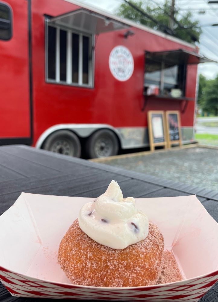 A made-to-order cider donut with a dollop of cannoli cream on top (and also inside) sits in a paper container on a table. In the background is a bright red food truck.