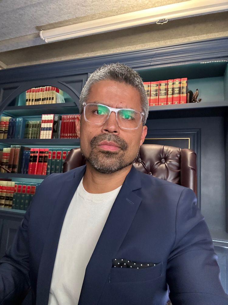 Cristian selfie. He’s sitting on a big leather chair with a shelf-full of casebooks and legal treatises behind him. He’s wearing a blue blazer, cream knit t-shirt, and a black pocket square with white dots.