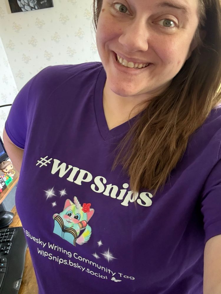 A selfie of Rose (a white woman with shoulder length brown hair) wearing a purple shirt with a caticorn illustration and the words #WIPSnips a Bluesky writing community tag. 