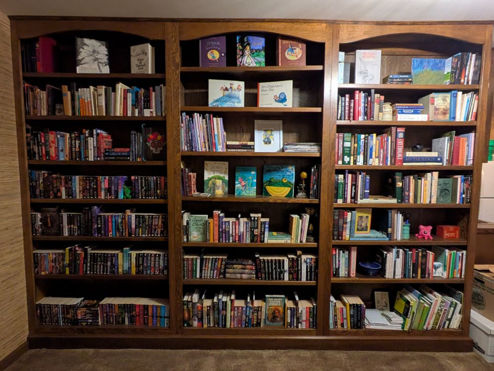 Wood built-in bookshelves full of books with display elements of objets d'art and pop-up books. From top left: Lit-fic, poetry, SF, PNR, children's books, textile craft and history, mystery, UF, fairy tales, mythology, folklore, philosophy, linguistics, writing craft and business, gardening.