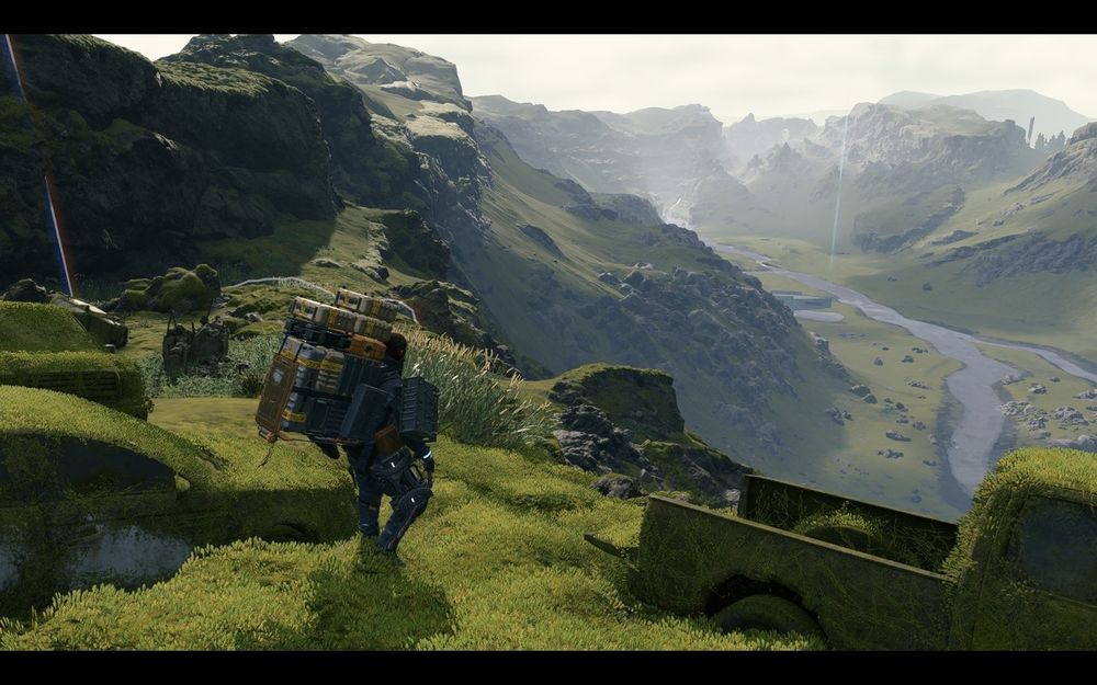 A person with a heavy backpack walks through grassy, mountainous terrain with rivers and moss-covered vehicles in the foreground.