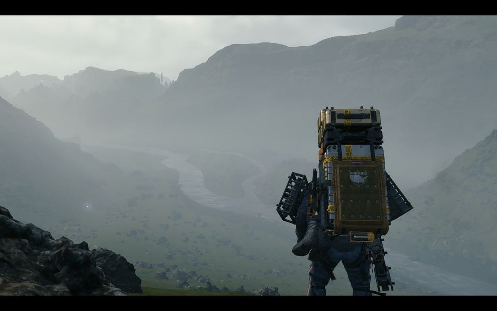A person wearing a heavy backpack stands on a rocky hill overlooking a foggy valley with a winding river and distant mountains.