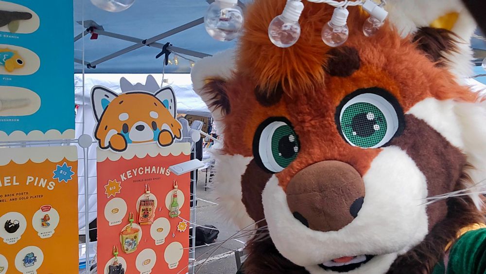 Burbley the red panda points out some red panda themed Keychains at a street vendor booth. 