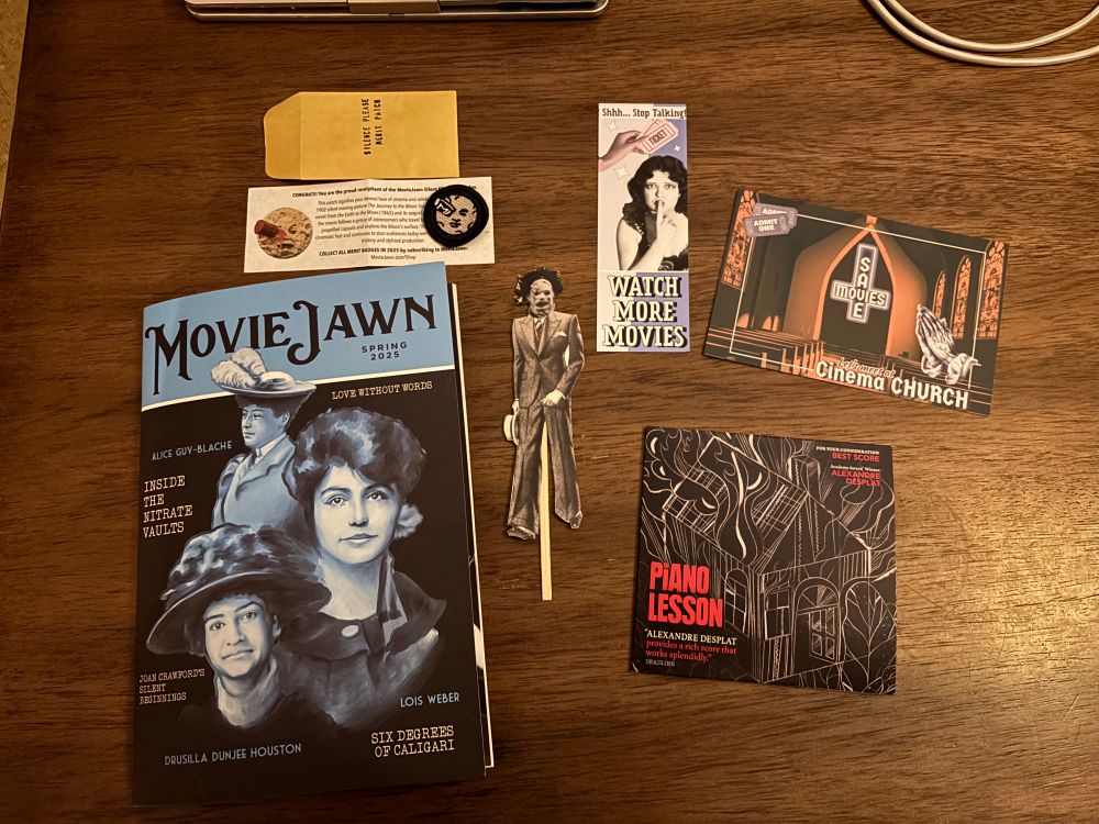 The image shows the contents of the latest MovieJawn mail drop, including a magazine in a blue and black color style showing visages of Alice Guy-Blanche, Lois Weber, and Drusilla Dunjee Houston, the copy of the score for “The Piano Lesson”, a bookmark which reads “Watch More Movies”, a patch, a postcard, and a black and white cutout photo of Leatherface dressed in 1920/30s garb.