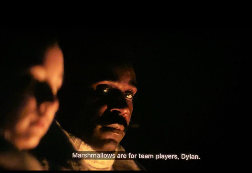 A man named Seth Milchick scolds his employee for acting out during a team building exercise. His assistant is seated next to him, slightly out of focus. Their faces are lit by campfire. Milchick looks terrifying yet calm.