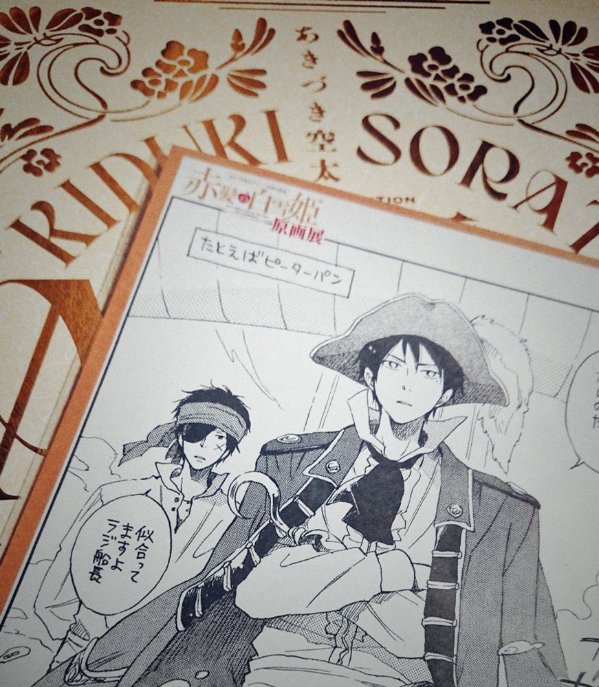白地に金の装飾がある原画展の画集と、その上に海賊風の衣装を着たラジの絵のカードが置かれているのを撮った写真。