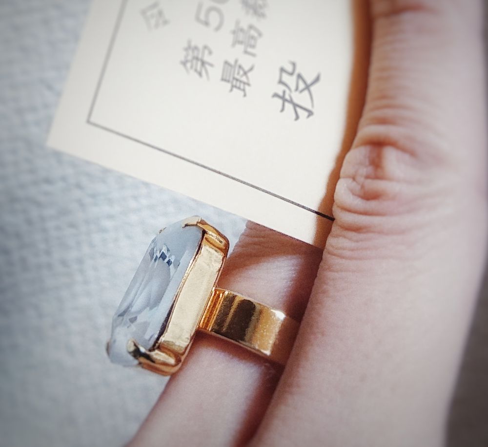 ひだりてのひとさしゆびとなかゆびで　とうひょうずみしょうめいしょお　はさんでいるしゃしん。　なかゆびに　おおつぶのとうめいな、　しかくいがらすのはまった　ごーるどのりんぐおつけている。　にちにちさんのりんぐです