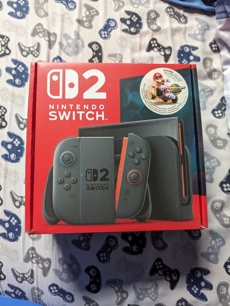 A brand-new Nintendo Switch 2 (+ Mario Kart World bundle) sits on a bed with white, video game controller-filled sheets.