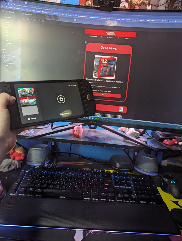 I'm holding my Switch 2 out in front of me in my left hand, above a desk with various knickknacks and a black keyboard. On my monitor, an email from Nintendo reads, "Good news! Nintendo Switch 2 System is waiting for you!"