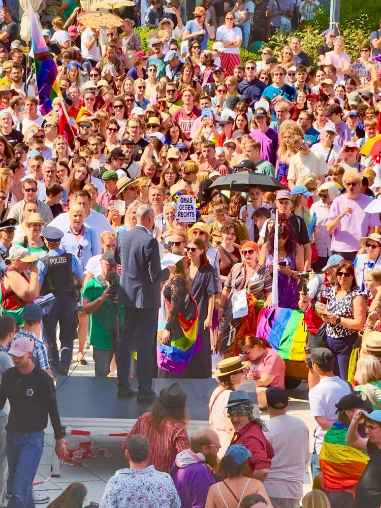 Das Bild zeigt eine große Menschenmenge auf einer Demonstration, wahrscheinlich eine LGBTQ+-Demonstration, mit einem Mann im Anzug, der eine Rede hält.


Detaillierte Beschreibung:

Das Bild zeigt eine große, dicht gedrängte Menschenmenge, die sich im Freien auf einem Platz oder einer Straße befindet. Die meisten Menschen tragen farbenfrohe Kleidung, und viele halten Regenbogenfahnen oder Schilder hoch.  Eine auffällige Regenbogenflagge ist im Vordergrund zu sehen, andere kleinere Regenbogenfahnen und -symbole sind in der Menge verteilt. Ein Schild mit der Aufschrift "OMAS GEGEN RECHTS" (Omas gegen Rechts) ist ebenfalls sichtbar, was auf einen politischen Kontext und einen Protest gegen rechte Ideologien hindeutet.


Im Zentrum des Bildes steht Karsten Vogt , Oberbürgermeister von Bautzen, im dunklen Anzug, der eine Rede hält.  Er steht auf einer kleinen, erhöhten Plattform. Um ihn herum stehen oder sitzen mehrere Personen, darunter eine Frau mit einer Regenbogenflagge.  Eine Person hält ein Mikrofon in Richtung des Mannes im Anzug.  Ein Polizist in Uniform ist ebenfalls sichtbar, was auf eine beaufsichtigte Versammlung hindeutet.


Die Atmosphäre ist lebendig und energiegeladen, mit einem Gefühl von Gemeinschaftsbewusstsein und politischer Aktivität. Die Sonne scheint hell, und der allgemeine Eindruck ist der einer friedlichen, aber leidenschaftlichen Demonstration. Viele Menschen tragen Sonnenbrillen, Hüte und andere Kopfbedeckungen. Die Menge besteht aus Menschen unterschiedlichen Alters und Geschlechts, was die Vielfalt der Teilnehmer unterstreicht. Der allgemeine Farbbereich des Bildes ist warm und hell, mit vielen bunten Kleidungstücken und Accessoires, die das Gefühl der Freude und des positiven Widerstands unterstreichen. Die allgemeine Stimmung ist positiv und hoffnungsvoll, trotz des politischen Kontextes des Protestes.
