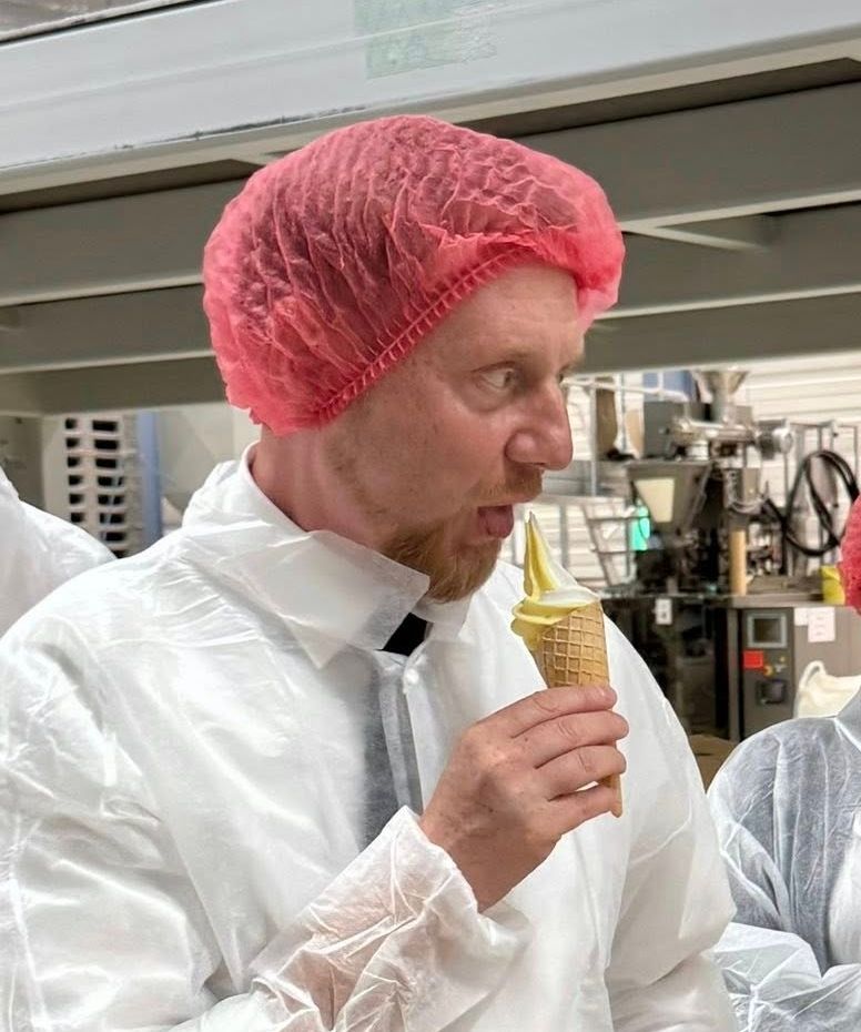 Das Bild zeigt den sächsischen Ministerpräsidenten Michael Kretschmer. Er trägt eine rosafarbene Haarnetzhaube, die sein kurzes, hellbraunes bis rötliches Haar bedeckt.  Sein Bart ist kurz und ebenfalls hellbraun bis rötlich.

Seine Gesichtszüge sind eher rundlich. Er hat hellblaue oder graublaue Augen, eine leicht breite Nase und einen geöffneten Mund, da er gerade von einem Softeis-Hörnchen isst. Sein Gesichtsausdruck ist konzentriert und genießerisch,  er scheint das Eis zu kosten.

Der Mann ist von mittelkräftigem Körperbau und steht aufrecht. Er trägt einen weißen, langärmligen Schutzkittel, darunter scheint ein dunkleres Hemd oder eine dunkle Krawatte zu erkennbar zu sein.  Die Schutzkleidung ist schlicht und ohne weitere Verzierungen.  Als Accessoire ist lediglich die Haarnetzhaube zu nennen.

Zusammenfassend lässt sich der Mann als Person beschreiben, die sich in einer Arbeitsumgebung befindet und scheinbar eine Pause einlegt, um ein Eis zu genießen. Die Kleidung deutet auf einen hygienisch sensiblen Arbeitsbereich hin.  Sein Ausdruck ist positiv und auf das Eis konzentriert.
