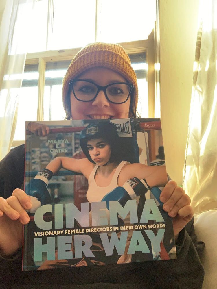 Cute writer wearing a yellow beanie and black-rimmed glasses holding up a book that she wrote called CINEMA HER WAY: VISIONARY FEMALE DIRECTORS IN THEIR OWN WORDS that has a photo of Michelle Rodriguez in Karyn Kusama's GIRLFIGHT on the cover