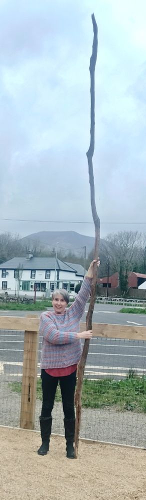 White person wearing jeans and a woolen jumper and a huge smile holding a gigantic stick - at least 15 foot high, no exaggeration in front of a mountain in the background