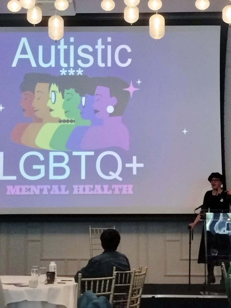 A white person wearing deal clothes, using a stick behind a podium in front of a large screen with text 'Autistic LGBTQ+ mental health' 