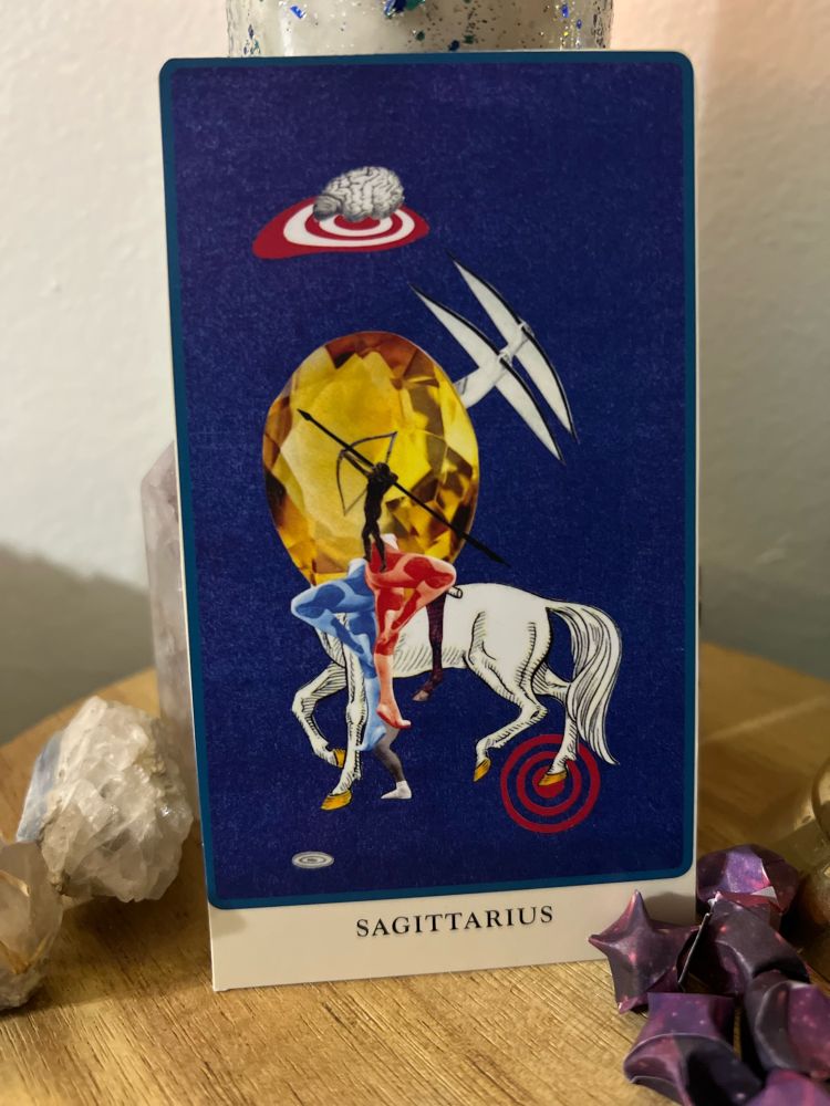 Sagittarius card from the Spolia Tarot deck - a collage of a white horse, yellow gemstone, archer silhouette, and bullseyes on a wooden platform with purple paper stars and various crystals 