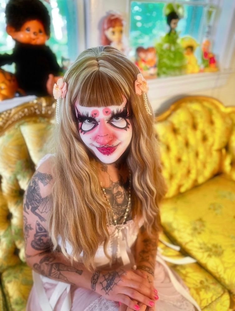 LEECH or Kitty in drag looking up sweetly at the camera. Wearing a blonde wig and graphic drag makeup. They are wearing a cute pink night gown and white vintage medical corset 