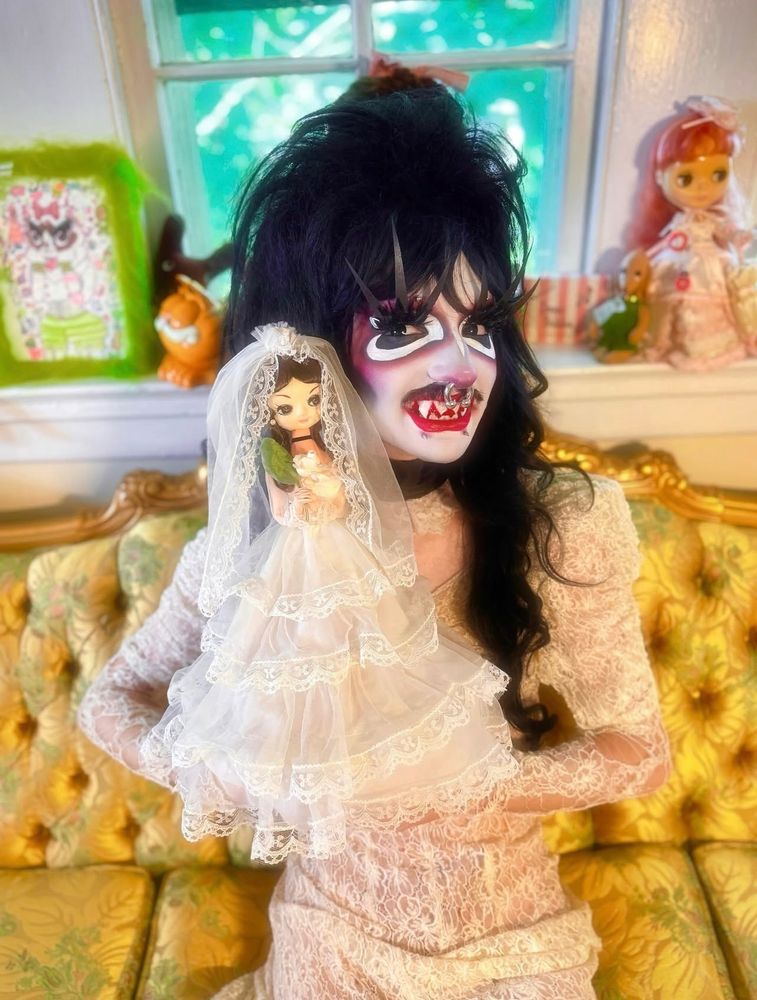 Piranha or Nicki in drag as a Victorian vampire, holding a vintage vampire doll wearing a similar outfit to them. They have a huge teased black wig on and graphic drag makeup and fangs. They are wearing a beautiful Victorian lace gown and smiling at the camera with their cute fangs popping out. 