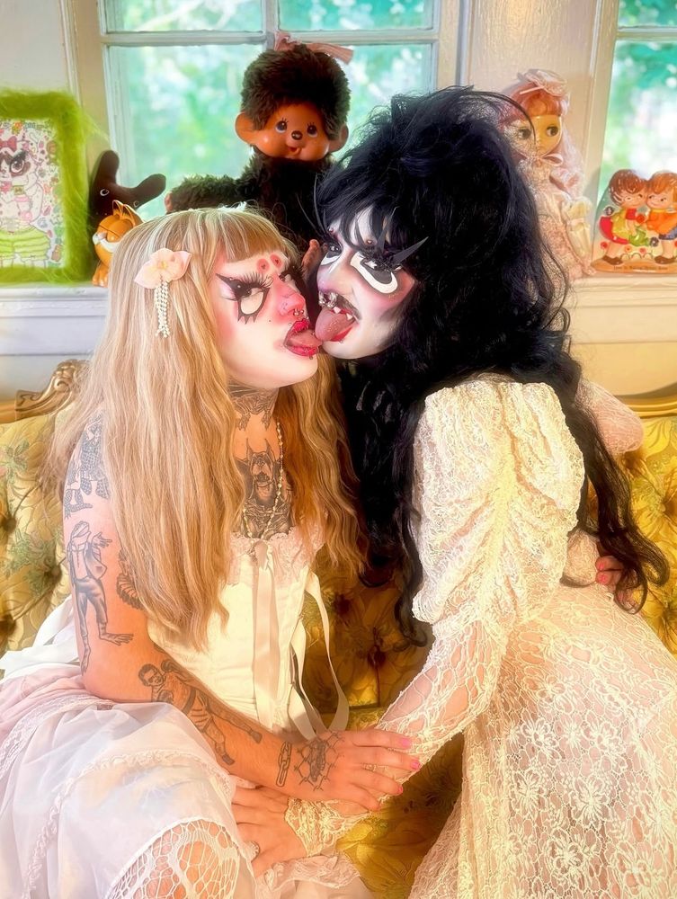 two drag queens in Victorian outfits, LEECH or Kitty is on the left and Piranha or Nicki is on the right. Piranha is a vampire and Leech is their familiar and they are holding each others arms and are touching tongues and looking super cute 
