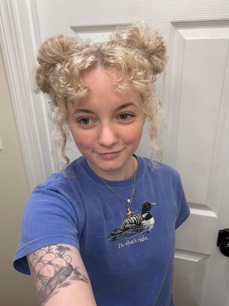 a selfie of a person with bleach-blonde hair, put up into two space buns. they have short curly bangs with some longer pieces out on the side