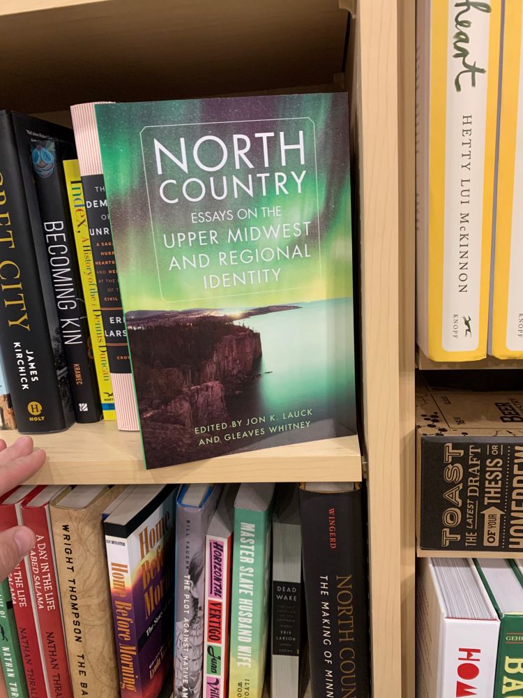 A book on a bookstore shelf titled "North Country: Essays on the Upper Midwest and Regional Identity," with a cover image of Palisade Head on Lake Superior under the Northern Lights.