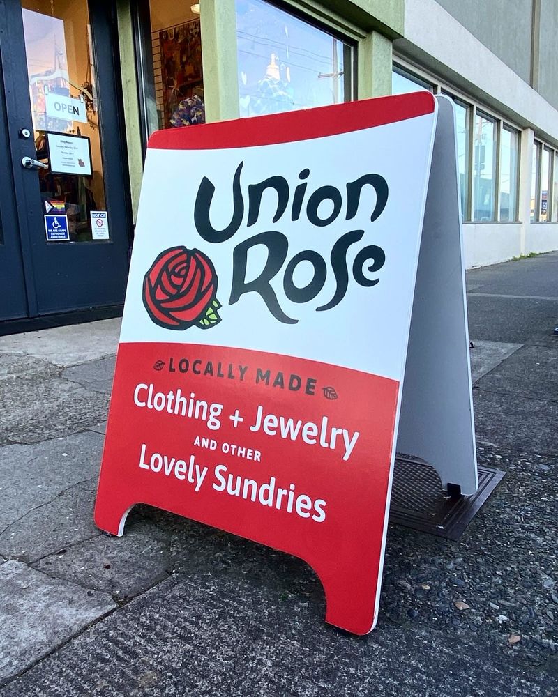 A red and white a board sits on the sidewalk. It features the logo, large on a white background, and within the red bottom half reads “locally made clothing and jewelry and other lovely sundries”