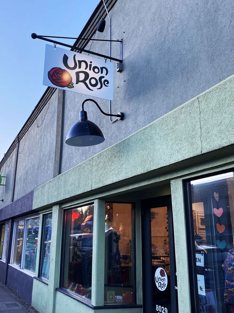 Exterior shot of the Union Rose shop on SE. Stark St. a blade sign showing the new logo with the rose oriented to the left of the typography is illuminated by a small spotlight, and a large round logo sticker is visible on the glass doorway.