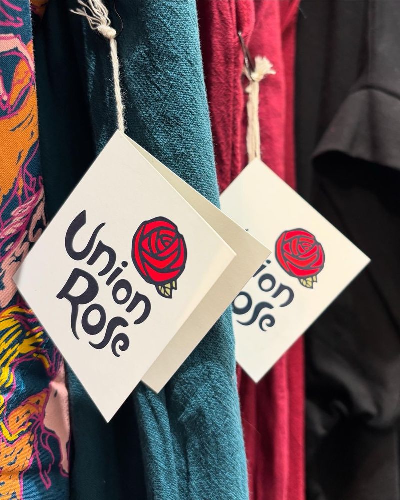 Folded garment logo cards hanging from a rack of beautifully colorful clothing. The featured logo is a craftsman style Rose, asymmetrically balanced over hand drawn typography reading “Union Rose”. Two shades of saturated red, two shades of sage green, and a dark dark gray.
