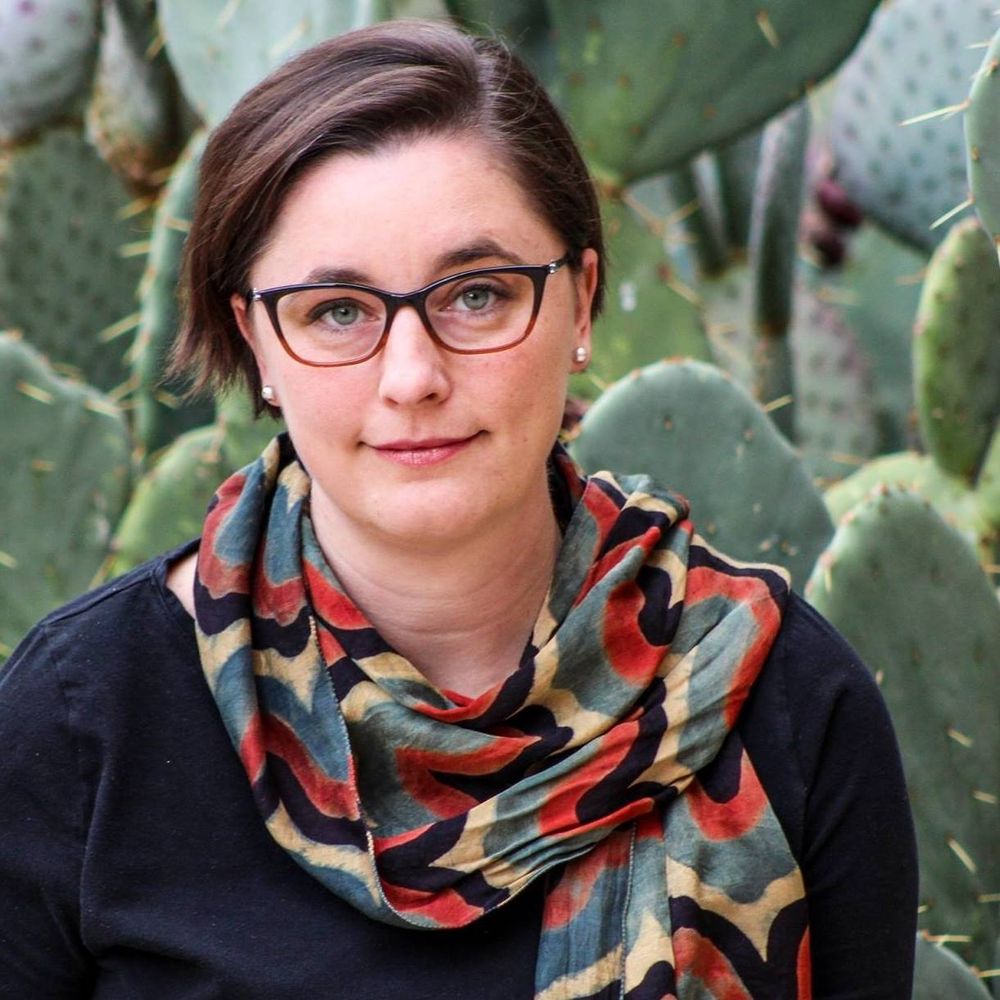 A Lady Professor with brown hair and glasses, wearing a scarf in red, green, yellow, & black, a gift from an incredible lactation  consultant, sitting in in front of cactus. 