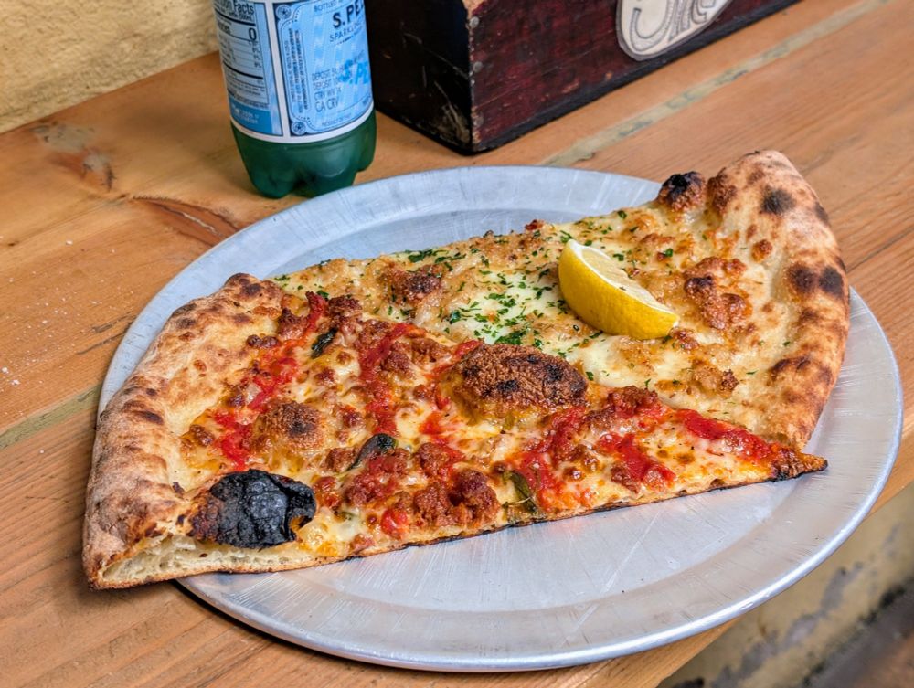 Two slices of thin, crispy pizza on a metal plate. One has a typical red sauce; the other is white with clams and a wedge of lemon on top.