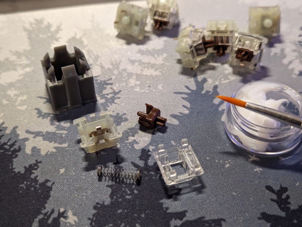 Cherry MX key switch in parts, with a switch opening tool, lube, and brush