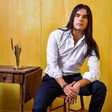 Native man with long black hair looking straight on in white dress shirt and navy dress pants sitting in from of yellow background next to old style desk