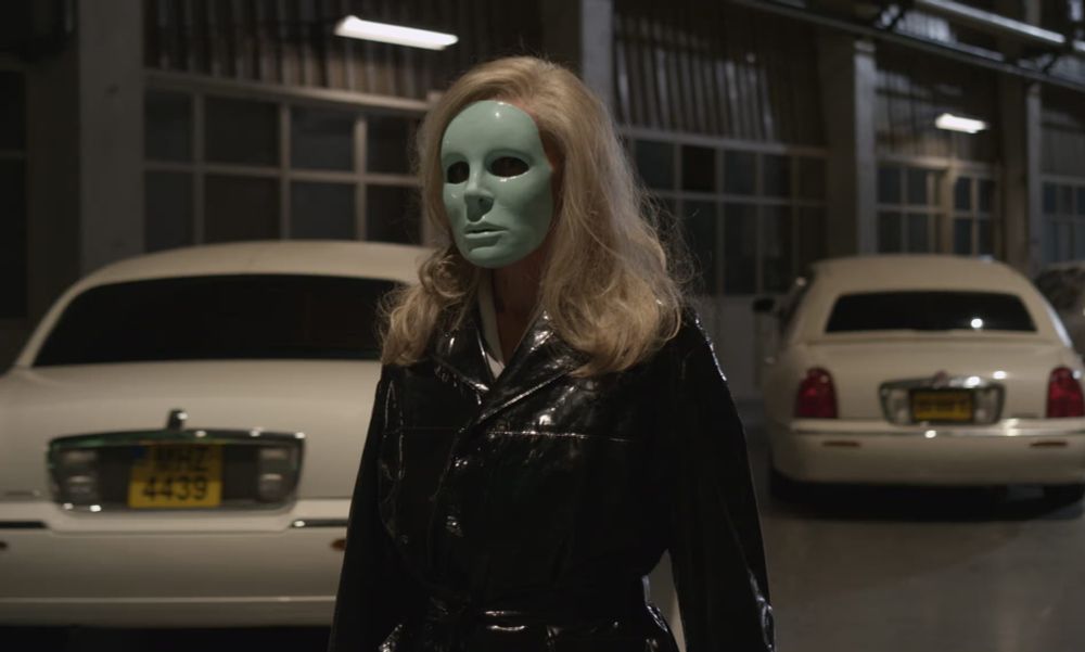 Screencap from HOLY MOTORS of Édith Scob wearing her EYES WITHOUT A FACE (1960) mask in a garage.