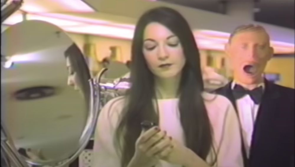 Still from Possibly In Michigan, depicting the protagonist partially reflected in the mirror, while The Man looks over her shoulder