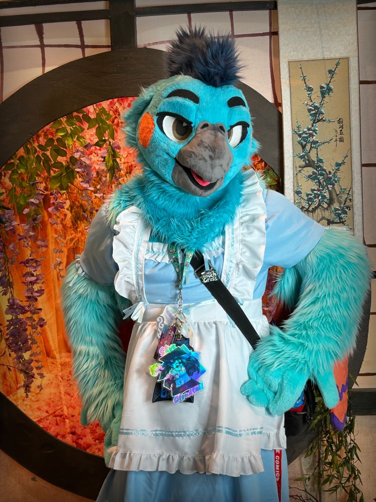 Blue cockatiel fursuiter wearing a maid outfit. They are posing with their hands on their hips in front of an Asian themed background.