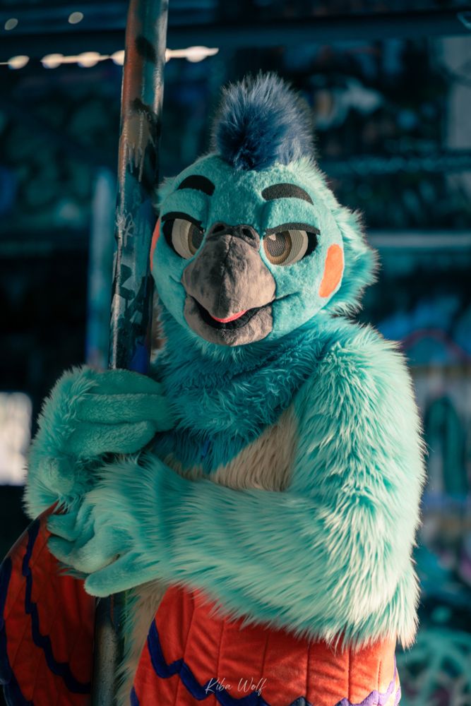 A blue cockatiel fursuiter holding onto a pole sticking out of the ground in a grafittied warehouse.