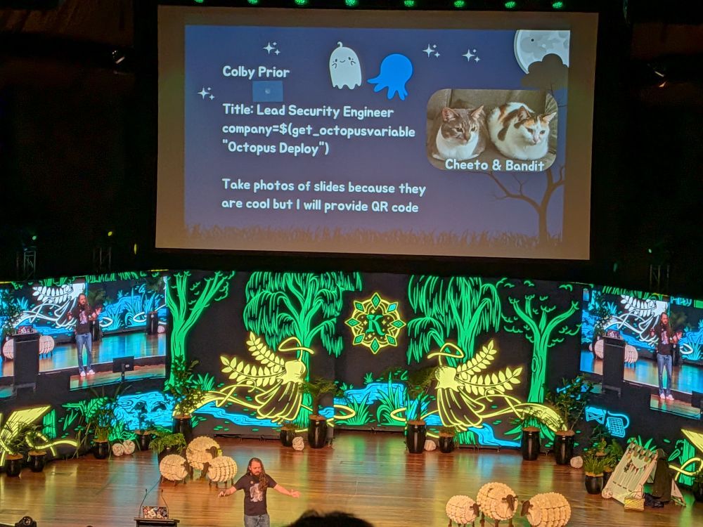 Kawaiicon stage, with Colby Prior standing front and center with his arms wide. Pretty coloured designs are behind him. Above him is a slideshow, showing his cats and employer, Octopus Deploy